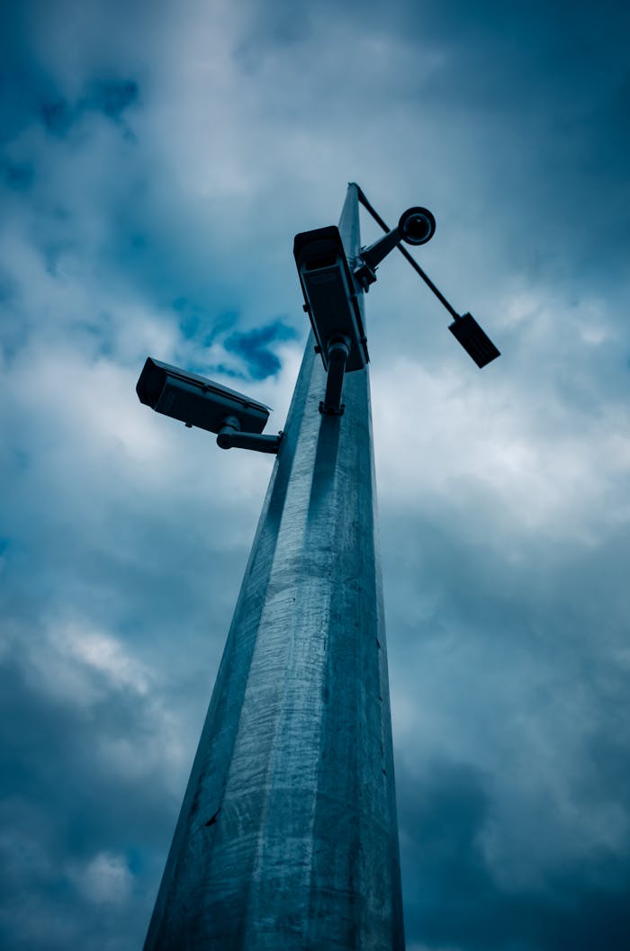creative Low-angle shot of CCTV cameras mounted on a tall pole against a cloudy sky.